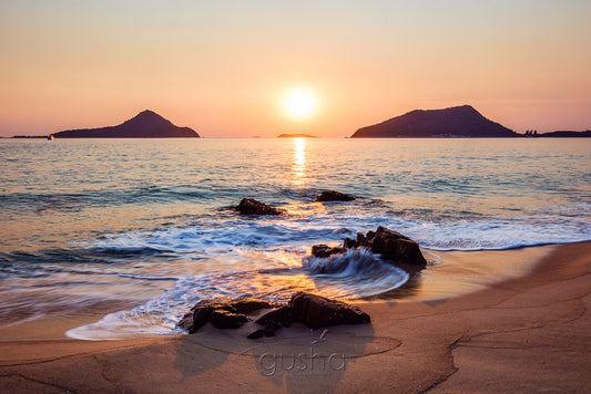 Captured from the sands of Shoal Bay, the sun rises between the heads of Port Stephens, Yacaaba Headland to the left and Tomaree Headland to the right.